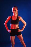 Standing wit,h the hands on waist. Beautiful muscular woman is indoors in the studio with neon lighting photo