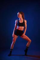 Front view, standing. Beautiful muscular woman is indoors in the studio with neon lighting photo
