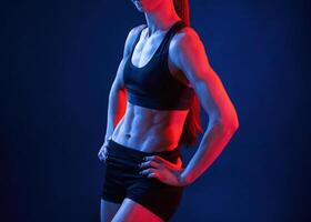 Standing with the hands on waist. Beautiful muscular woman is indoors in the studio with neon lighting photo