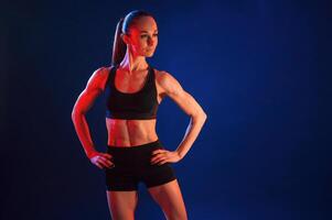 Hands are on the waist, standing. Beautiful muscular, woman is indoors in the studio with neon lighting photo