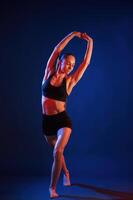 Sportive exercises, standing and stretching the hands. Beautiful muscular woman is indoors in the studio with neon lighting photo