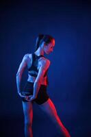 Back view. Warm up, stretching exercises. Beautiful muscular woman is indoors in the studio with neon lighting photo