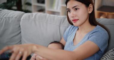 Selective focus shot, A beautiful mother gently holds her little son in her arms, feeds him milk with a bottle, and uses the remote control to select channels. photo