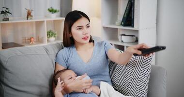 Portrait of A beautiful mother gently holds her little son in her arms, feeds him milk with a bottle, and uses the remote control to select channels photo