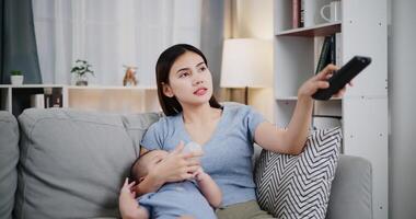 Portrait of A beautiful mother gently holds her little son in her arms, feeds him milk with a bottle, and uses the remote control to select channels. photo