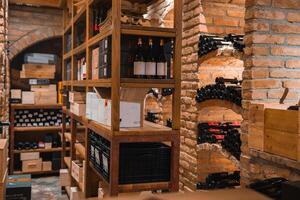 A well organized wine cellar with brick walls, arched alcoves, and wooden shelves stocked with wine bottles, crates, and boxes in warm lighting. photo
