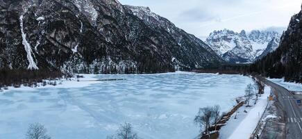 A frozen lake in the Alps surrounded by snow covered peaks, a winding road with sparse trees, and rugged mountain slopes in the background. photo