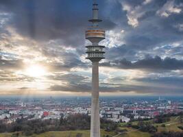 The Olympiaturm in Munich, Germany, rises against a sunset sky, with its observation deck visible. The cityscape below features diverse architecture and green spaces. photo