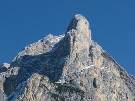 A rugged mountain peak with sharp rock formations and snow patches, set against a clear blue sky. Greenery accents the lower slopes in an alpine setting. photo
