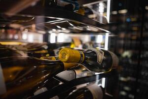 Horizontal wine bottles with yellow caps and detailed labels in a well lit, modern wine cellar with reflective surfaces and blurred racks. photo