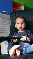 Curious child focused on the TV remote in his hands. Kid sits in big leather chair turning remote control in hands. Low angle view. Vertical video