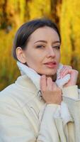 Caucasian brunette holding by the jacket collar stands in the rays of setting sun. Young woman spending peaceful time at river bank in autumn. Vertical video