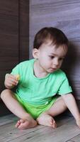 Curious toddler boy sits on the floor holding some food in his hand. Cute baby stands up and approaches camera. Low angle view. Vertical video