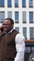 Portrait of happy african american man in city. Smiling black man in stylish glasses talking the phone while standing outdoors. Businessman with a phone looks at camera. Vertical video