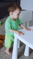 Pretty little toddler is in his room. Child wants to sit at the table with a range of bright toys on. View from top. Vertical video