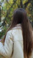 Attractive brunette walking by the autumn nature with phone in hand. Lady walking away from camera, turns for a moment smiling and turns around again. Vertical video