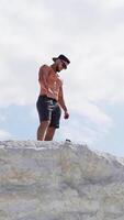 Shirtless athlete in cap and glasses on mountain. View from below on a sportsman standing on the top of white hill. Vertical video
