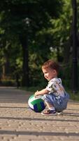 Little boy squats near the ball and pushes it to roll. Kid stands up and walks opposite side by the paved road. Vertical video