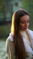 Relaxed woman leaned on the handrail of the bridge. Brunette lady using her phone near the river in autumn. Blurred backdrop. Vertical video