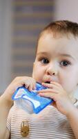 Lovely Caucasian toddler holds a pack of fruit puree in his hands. Baby tries to get some food from the pack with his finger. Close up. Blurred backdrop. Vertical video