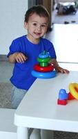 alegre caucasiano bebê Garoto tocam às lar. sorridente criança montagem a brinquedo pirâmide em a branco escrivaninha. topo visualizar. vertical video