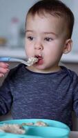 Little Caucasian toddler holding a fork in hand while mom feeds him from spoon. Caring mother takes the bits of food from kid's face with a spoon. Vertical video