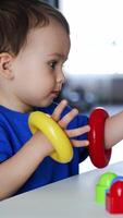 Charming cute Caucasian baby boy sits at the desk playing with pyramid. Toddler puts on the colorful rings on hands and clapping hands cheerfully. Vertical video