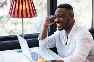 A smiling man sits at a table with a laptop photo