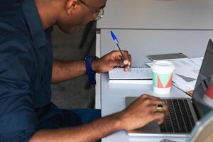 A man is writing on a notebook while sitting at a desk photo