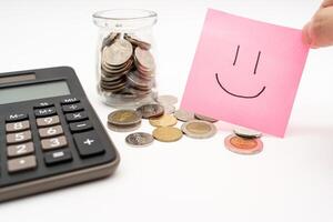 Saving money concept with Smiling faces drawn on pink sticky note. Commitment to achieving financial goals. isolated on white background. Thai Baht Coins spilled out with Calculator. photo