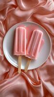 Two pink popsicles resting on a white plate atop soft, draped pink fabric creating a visually appealing and refreshing dessert display photo