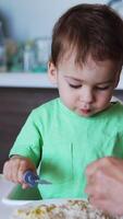Adorable toddler boy sits at little table with plate of porridge in front of him. Cute baby uses fork to eat. Blurred backdrop. Vertical video