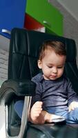 Beautiful toddler swinging in the big leather chair. Child plays with TV remote and saying something. Low angle view. Vertical video