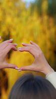 Unknown brunette outdoors holds up her hands to make the heart sign. Beautiful sunny autumn colorful trees at backdrop in blur. Vertical video