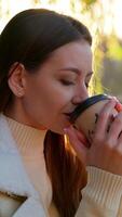 Enjoying hot coffee from a paper cup near the river. Long-haired attractive lady sitting under the tree in the rays of bright autumn sun. Blurred backdrop. Vertical video