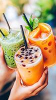Refreshing drinks in colorful glasses held by three hands, featuring iced bubble milk tea, green tea, and orange juice, perfect for sunny day photo