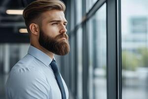 Young man with beard looking thoughtfully out of a large window in a modern office setting photo