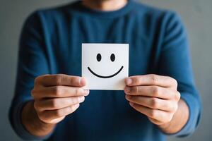 A person holds a smiling face on a note, conveying positivity and joy against a neutral background. photo