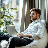Balancing Freedom and Flexibility with Remote Work. A man in a white shirt works on a laptop while relaxing in a cozy chair, surrounded by indoor plants and natural light streaming through a window. photo