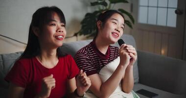 Handheld high angle view shot,Two Asian women sitting on a sofa happily singing karaoke with smiles and enjoying a fun time together at home. Share good times video
