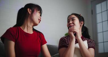 Handheld low angle view shot, Two Asian women sitting on a sofa happily singing karaoke with smiles and enjoy a fun time together at home. Share good times video