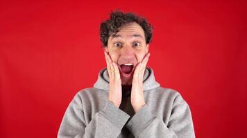 large close up portrait of young curly haired man, joyfully surprised, holding his face with his hands and mouth open, pronouncing word wow on studio red background. Concept - discounts, Black Friday. video