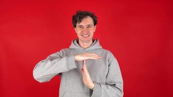 young adult curly haired brunette man holds his hands in front of shows sign timeout gesture. smiling, wearing grey hoodie. isolated on red studio background. Concept - pause, break, rest, vacation. video