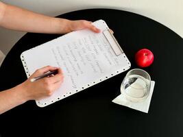 Female writing on clipboard with notes next to apple and glass on round black table photo