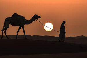 A man walking a camel in the desert at sunset photo