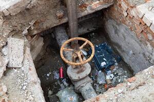 A rusty valve surrounded by trash sits in a decaying, dirty pit in an urban environment. photo