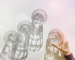 Transparent Water Glasses, hand holding one, shadows at sunlight on white, pastel color photo minimal style, aesthetic top view shiny cups sparkling at natural light, summer drink, composition