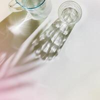 Transparent Water Glasses and water jug with palm leaf shadows at sunlight on white, pastel color photo minimal style, aesthetic top view shiny cups sparkling at natural light, summer drink