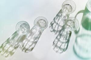 Water Glasses and water jug at Shadows and sunlight Reflection on white background, colored photo minimal style, aesthetic summer still life, refraction throug crystal shine cups, caustics