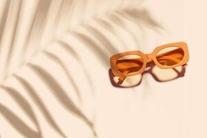 Trendy sunglasses with orange Frame on neutral beige background, copyspace, beach vibes, summertime mood concept, palm leaf shadow, summer fashion glasses. Minimal top view of eyeglasses photo
